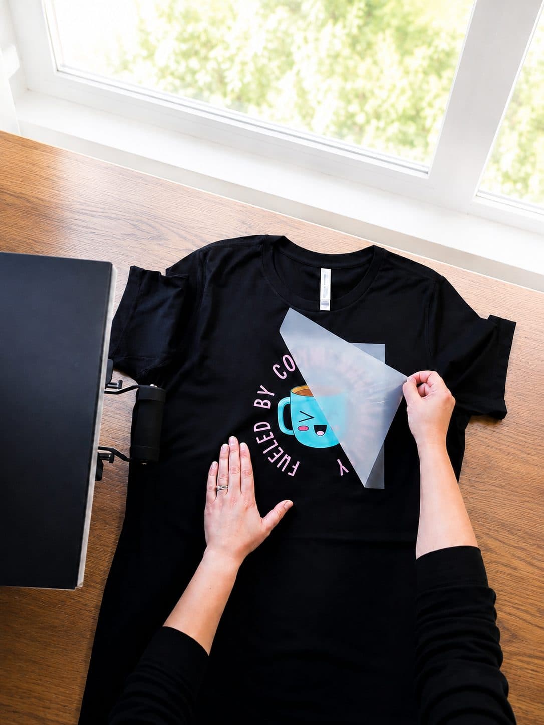 Hands peeling a DTF transfer with a coffee-mug design onto a black tee