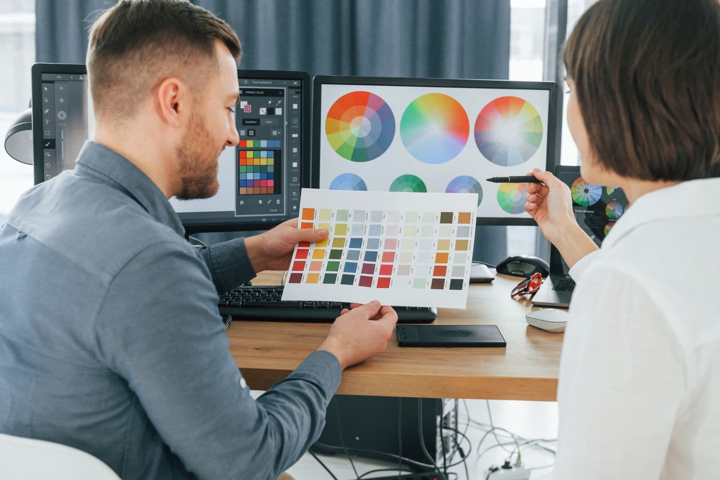 Prepress designer comparing a printed color swatch chart with the color wheels on screen