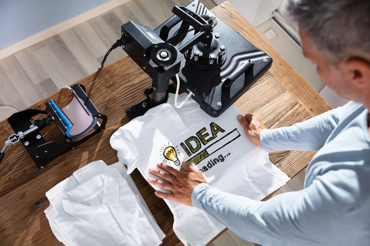 Print-shop operator lifting an "IDEA loading" transfer off a white tee at the heat press